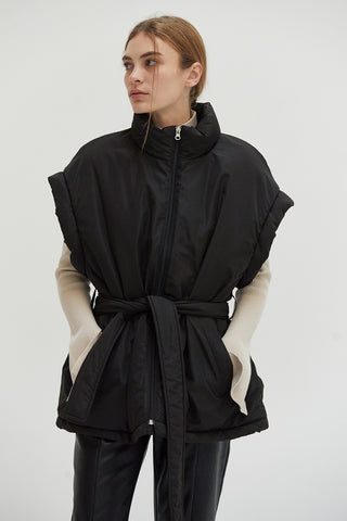 A woman wearing a black mock neck puffer vest with side pockets and detachable self belt. 